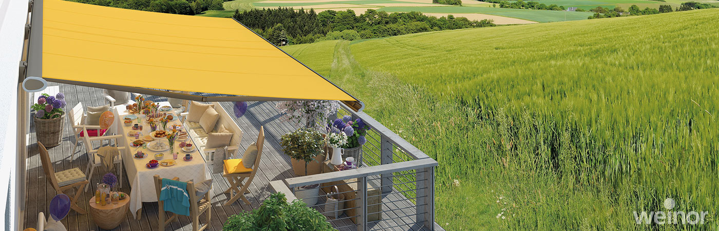 Vogelperspektive auf einen Balkon mit Blick auf ein grünes Feld. Eine gelbe Markise ist ausgefahren und bietet Sonnenschutz.